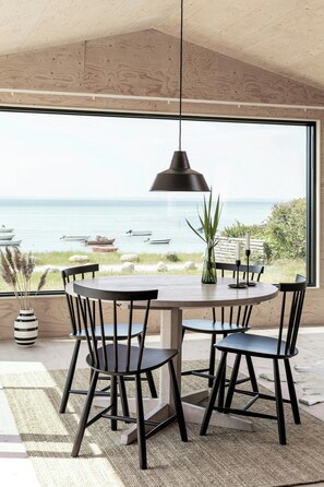 Uma área de refeições com vista para o mar, apresentando uma mesa redonda, cadeiras pretas, uma luminária pendente e um vaso com planta.