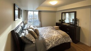 A bedroom with a bed, a dresser, a mirror, a window with curtains, and a ceiling light. - Times Square Next To Rogers Place #205 (EDMONTON)