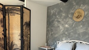 Quarto com cama, almofadas, mesa de cabeceira, chapéu fixado na parede e um biombo com palmeiras.