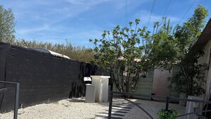 Uma entrada de cascalho com um portão de metal, uma lixeira branca e uma árvore em um bairro residencial.