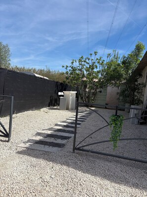 Une allée gravillonnée avec un portail en métal, une poubelle blanche et un arbre, le tout situé dans un quartier résidentiel.