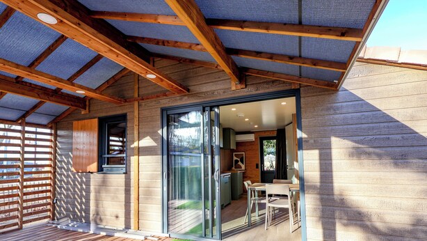 Eine Holzhütte mit überdachter Terrasse, einer Glasgleittür zu einem Essbereich und einer sichtbaren Küche.