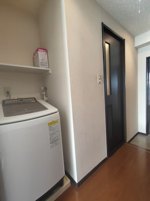 A laundry area with a washing machine, a shelf with a box, and a door leading to another room. - 10 min walk from JR Namba station and Subway/Hanshin Sakuragawa station. 4 min from JR Ashiwarabashi station and supermarket. 1 room on 1 floor, max 4 people, elevator, convenient to Namba, Dotonbori and Shinsaibashi. (大阪市)