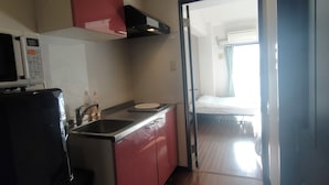 Eine kleine Küche mit Mikrowelle, Spüle und roten Schränken. Ein Durchgang führt in einen Schlafbereich mit Bett und Fenster.
