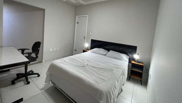 A bedroom with a bed, a desk, a chair, and a nightstand with a lamp. - 474flats e Suítes Standart  (Ipatinga)