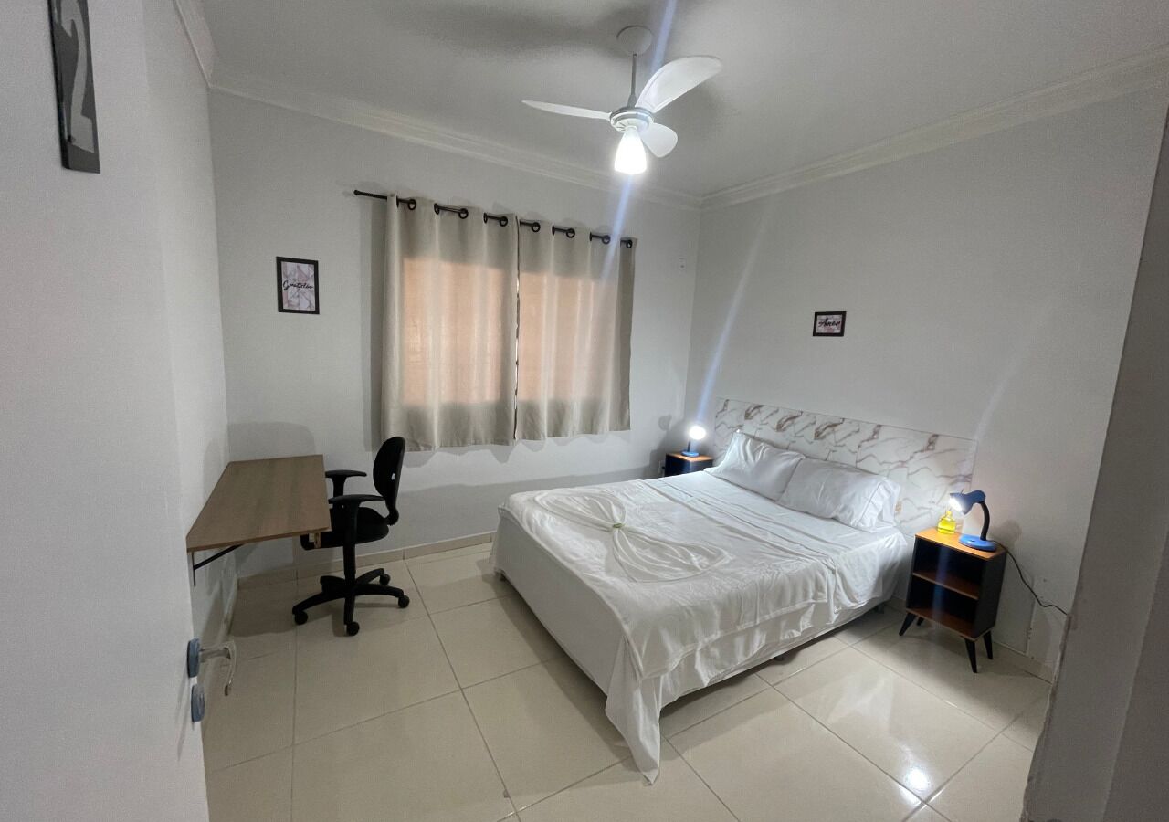 A bedroom with a bed, a desk, a chair, a nightstand, a ceiling fan, and a window with curtains.