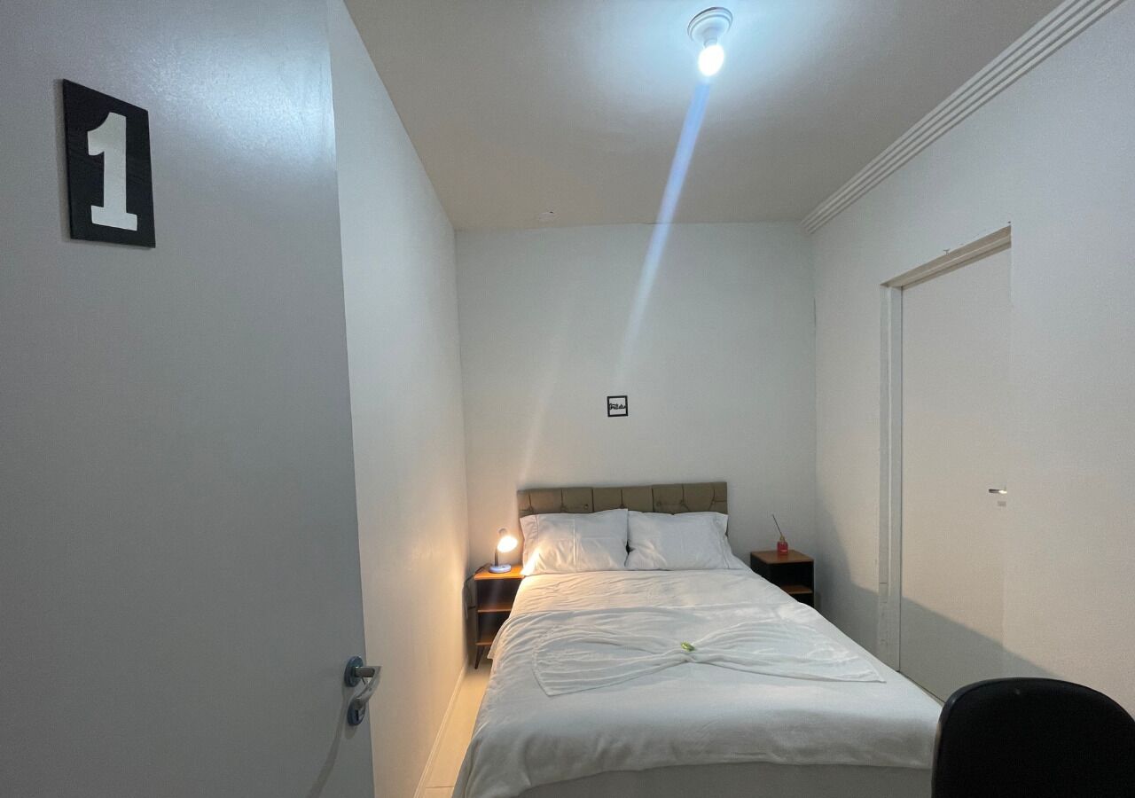 A single bed with a headboard, a bedside table with a lamp, a door with a number sign, and a ceiling light fixture.