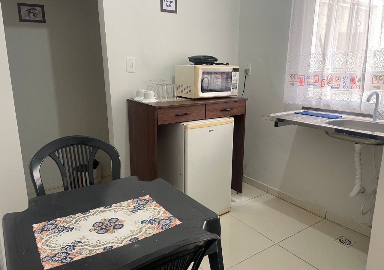 A compact kitchen with a microwave, refrigerator, and a small sink. There is a dining table with a patterned tablecloth and two chairs.