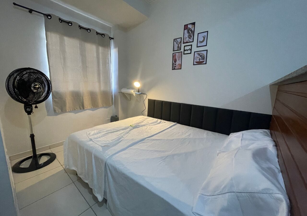 A bedroom with a bed, a standing fan, a bedside lamp, a window with curtains, and wall art.