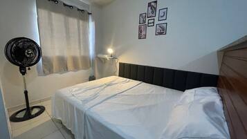 A bedroom with a bed, a standing fan, a bedside lamp, a window with curtains, and wall art.