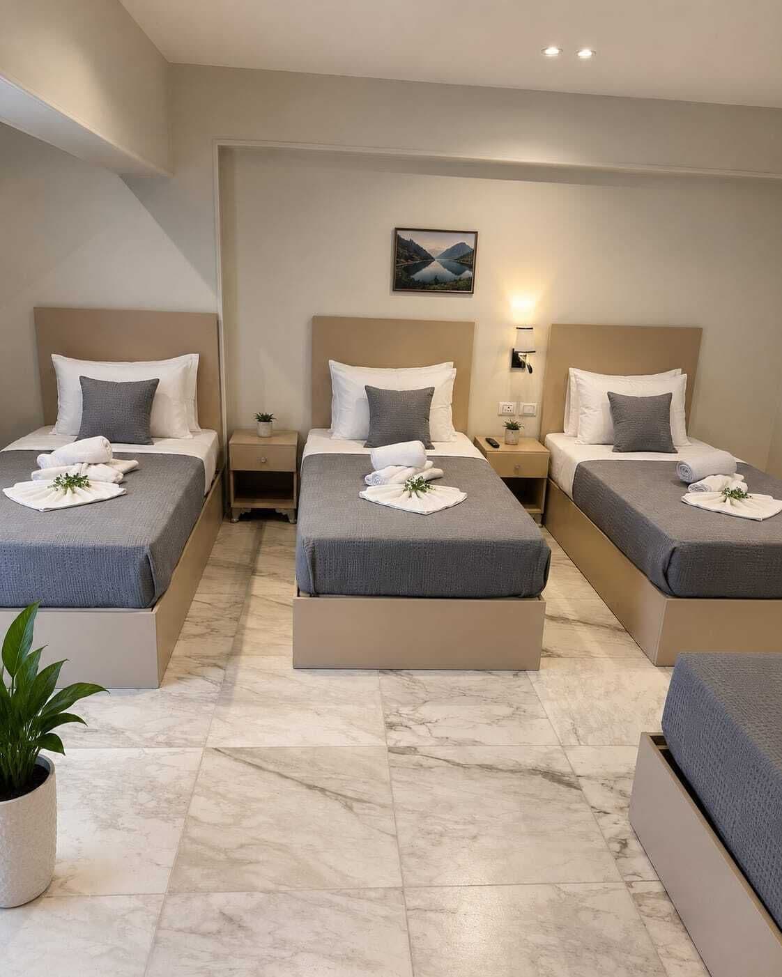 Habitación de hotel con tres camas, cada una con colcha gris y almohadas blancas, una mesita de noche con lámpara y un cuadro en la pared.