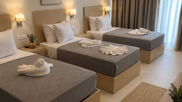 Hotellihuoneessa on kolme sänkyä, joissa kaikissa on pääty ja yöpöydällä lamput. Huoneessa on suuri ikkuna, jossa on verhot, seinällä on kehystetty taulu ja yöpöydällä pieni kasvi.