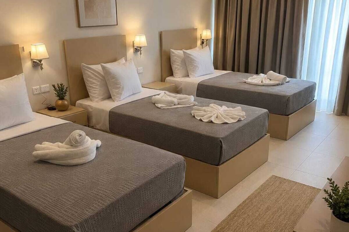 A hotel room with three beds, each with a headboard and bedside lamps. There is a large window with curtains, a framed picture on the wall, and a small plant on the nightstand.