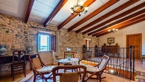 Ein rustikales Interieur mit Steinsäulen, Holzbalken, einem Essbereich mit Stühlen und Tisch sowie einer Treppe mit Geländer.