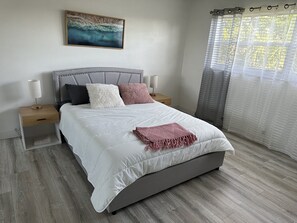 Ein ordentlich eingerichtetes Schlafzimmer mit Bett, Nachttischen, einem Tischchen, einem Gemälde und einem Fenster mit Vorhängen.