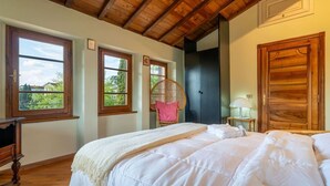 A bedroom with two beds, wooden beams on the ceiling, a wicker chair, and a wardrobe. - 300-square-meter villa · 6 bedrooms & 5 bathrooms · Cool in the summer (Bibbiena)