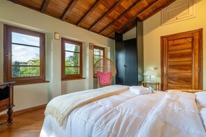 A bedroom with two beds, wooden beams on the ceiling, a wicker chair, and a wardrobe.