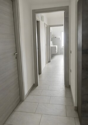Un corridoio con pavimento piastrellato chiaro e pareti bianche, che conduce a un'area bagno con lavandino e specchio.