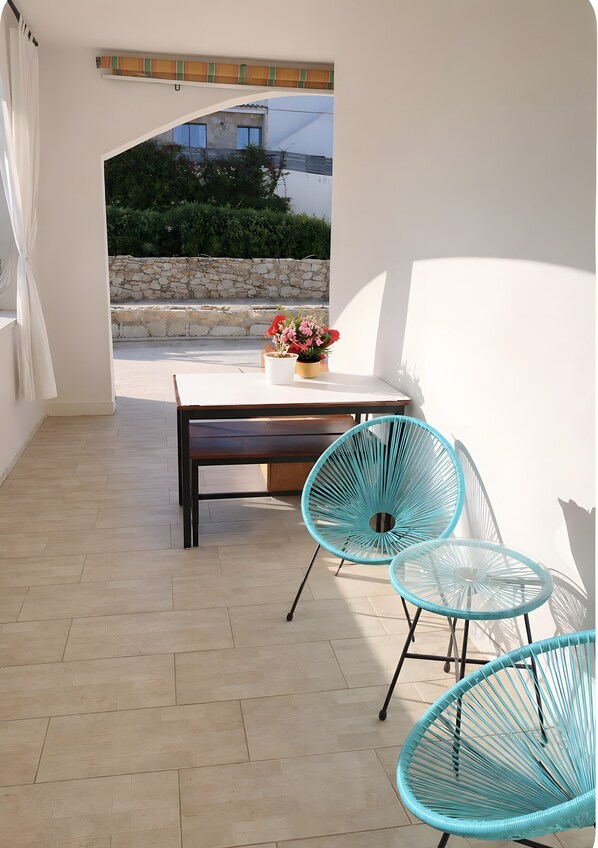 A patio with a table and chairs, a potted plant, and a view of greenery and a building.