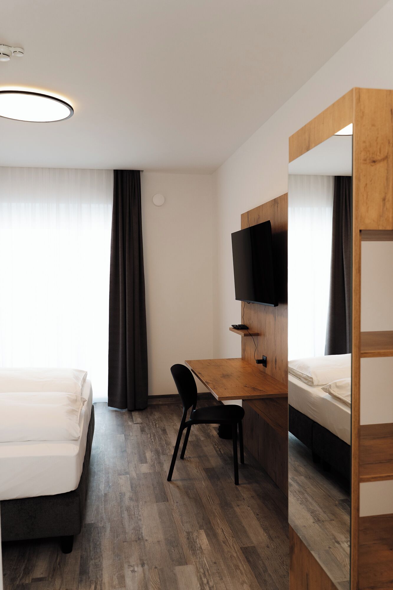 Habitación de hotel con cama, escritorio con silla, televisor montado en la pared y literas de madera.
