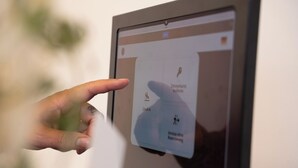 Una mano interactuando con una interfaz digital, posiblemente un quiosco de registro, con iconos para el registro, selección de habitación y un mapa.