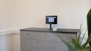 Un mostrador de recepción con un monitor de computadora, un detector de humo en el techo y una planta al frente.