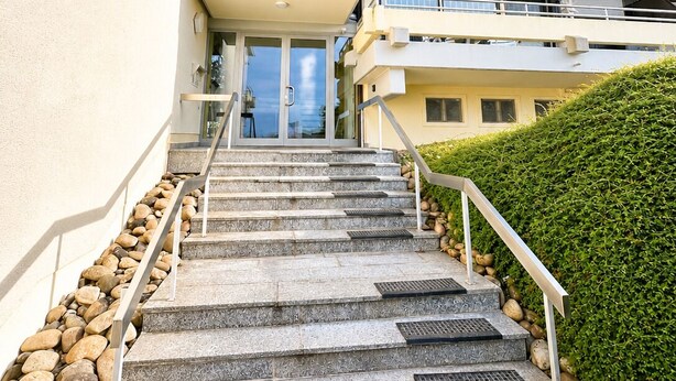 A multi-story building with a staircase leading to the entrance, a flat roof, and a hedge on the right.