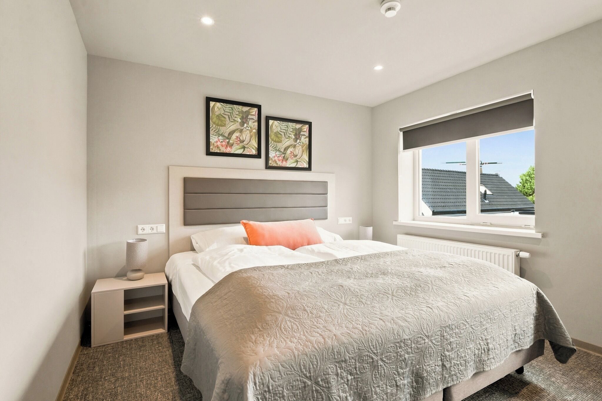 Een slaapkamer met een bed, nachtkastjes, een radiator, een raam met uitzicht en twee ingelijste foto's aan de muur.