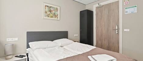 A neatly made bed with a quilted cover, a bedside table with a lamp and remote controls, a door with a wooden panel, and a wall-mounted painting.