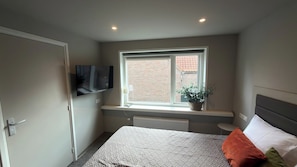 A bedroom with a bed, a TV mounted on the wall, a window with a view of a brick building, and a radiator under the window.