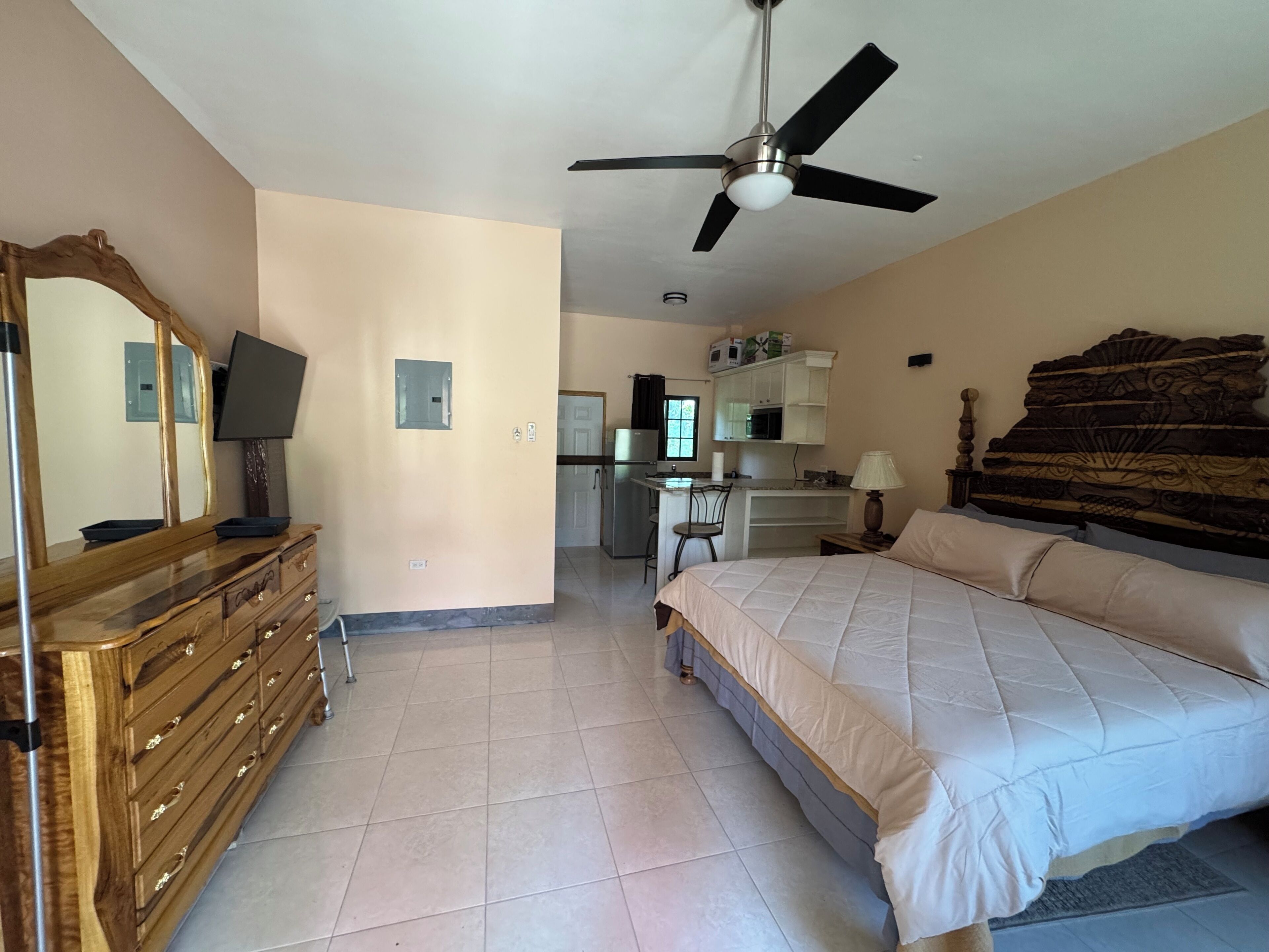 Ein Schlafzimmer mit Bett, Schminktisch, Deckenventilator und einer kleinen Küche im Hintergrund.