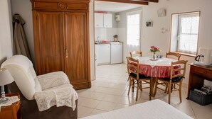 Een kamer met een bed, een stoel, een tafel met een vaas, een houten garderobe, een keuken op de achtergrond en een raam met uitzicht.