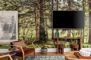 A living room with a forest-themed wallpaper, a large flat-screen TV, a brown leather sofa, a wooden coffee table, and a potted plant. - 2 Units, Sleeps 24  Nashville's Best Location (Nashville)
