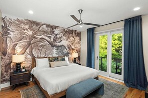 A bedroom with a large bed, a ceiling fan, a nightstand with a lamp, a bench, and a view of greenery through the window. - 2 Units, Sleeps 24  Nashville's Best Location (Nashville)