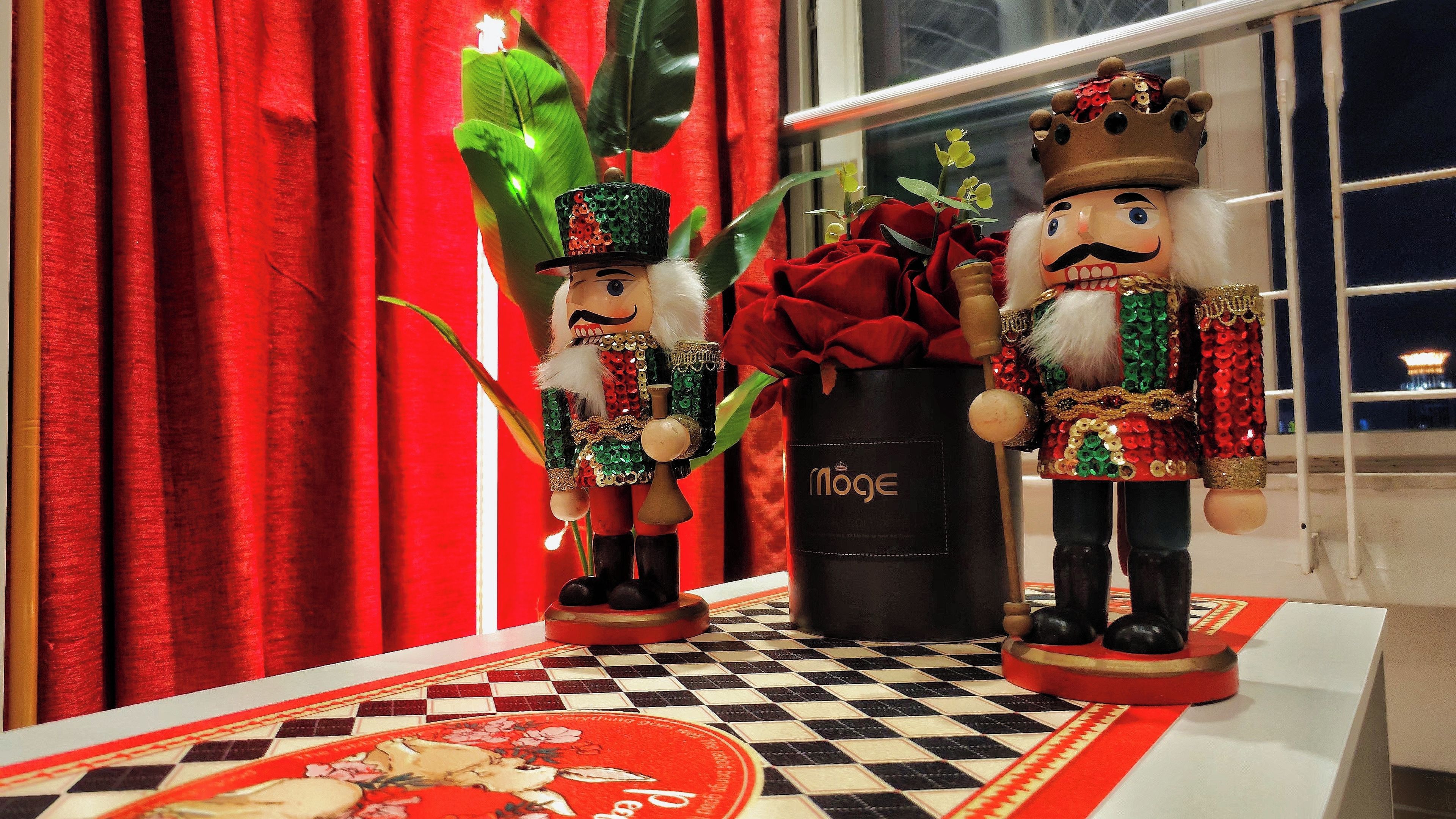 Two nutcracker figurines on a checkered tablecloth with a red and white pattern, a red curtain, and a plant in the background.