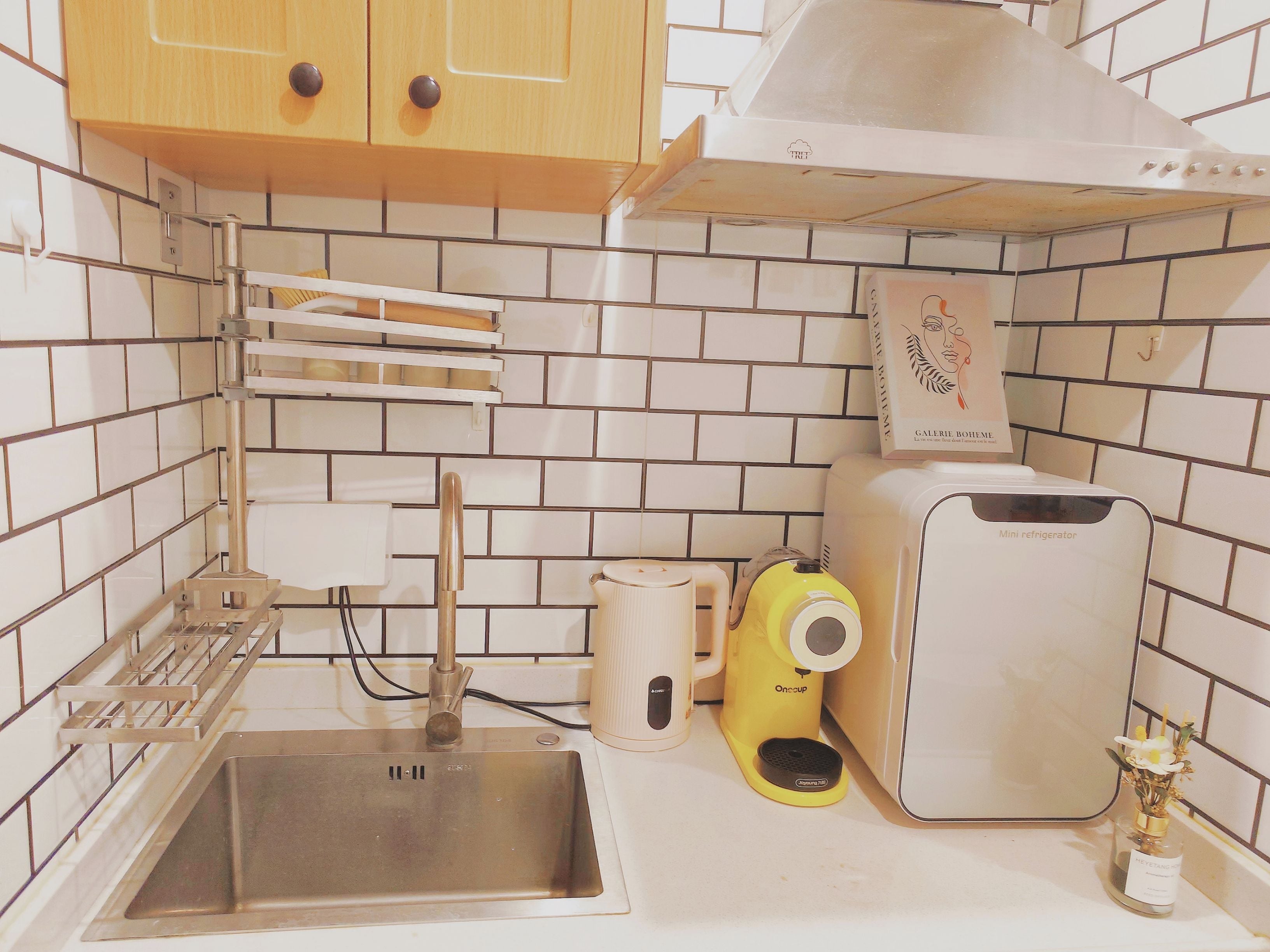 A kitchen with a stainless steel sink, a yellow Onecup coffee maker, a mini refrigerator, and a cabinet with a white handle.