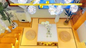 Um quarto com piso de madeira, uma mesa com um objeto decorativo no centro, um ventilador de teto e uma janela com cortinas.