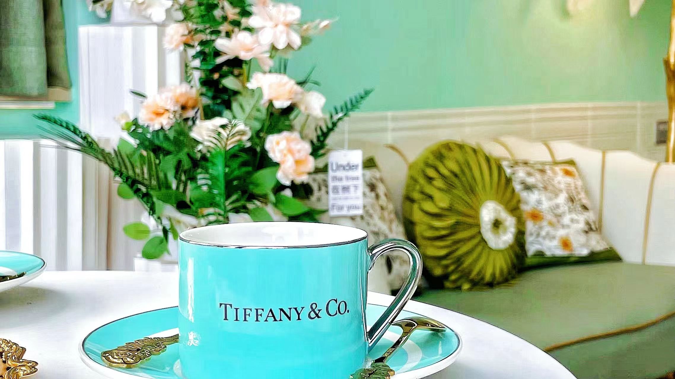 Une tasse Tiffany & Co. posée sur une soucoupe, accompagnée d’une cuillère dorée, d’un arrangement floral vert et d’un canapé vert agrémenté d’un coussin à motifs, dans une pièce où l’on aperçoit un radiateur blanc et un mur vert.