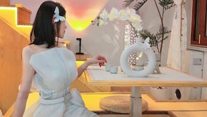 Uma mulher de vestido branco sentada à mesa com um anel decorativo branco, um pequeno bule e duas chávenas.
