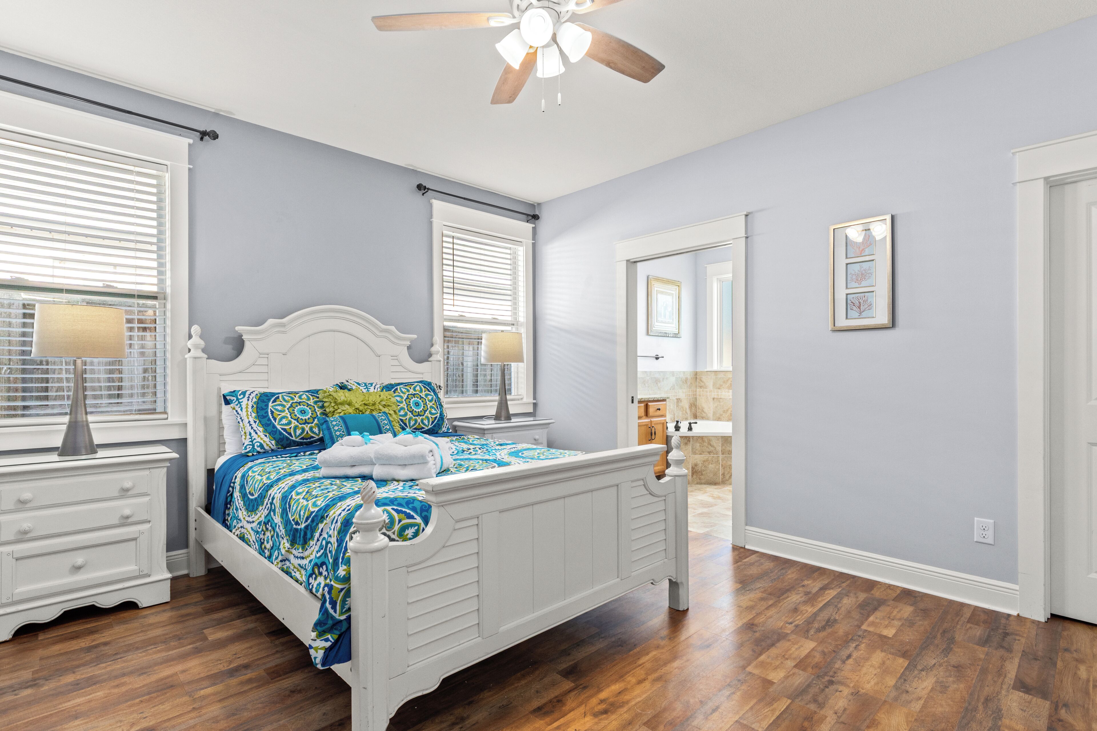 A bedroom with a white bed, wooden flooring, a ceiling fan, and a window with blinds.