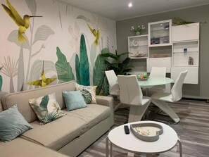 Ein Wohnzimmer mit einer Couch, Sesseln, einem Couchtisch und einem Esstisch. An der Wand befindet sich ein Wandgemälde und ein Bücherregal.