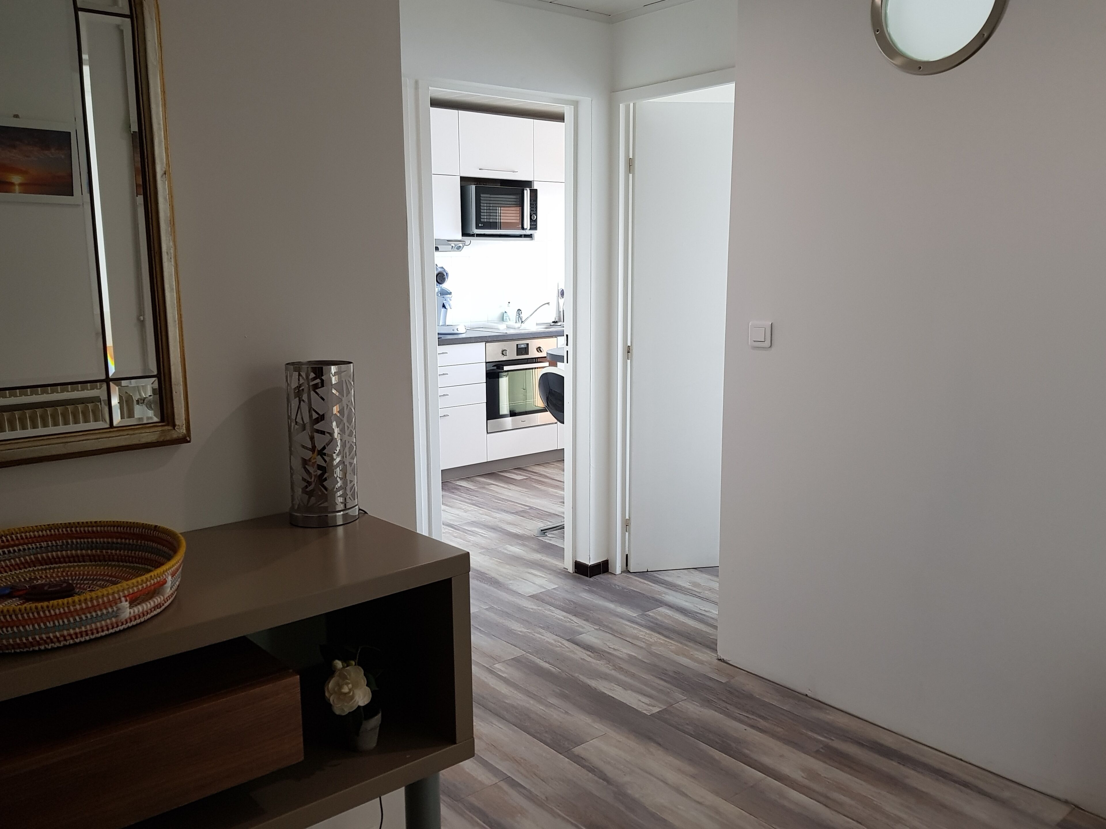 Un pasillo que conduce a una cocina con gabinetes blancos, un microondas y un horno. Se observa una mesa consola de madera con un florero y una cesta.