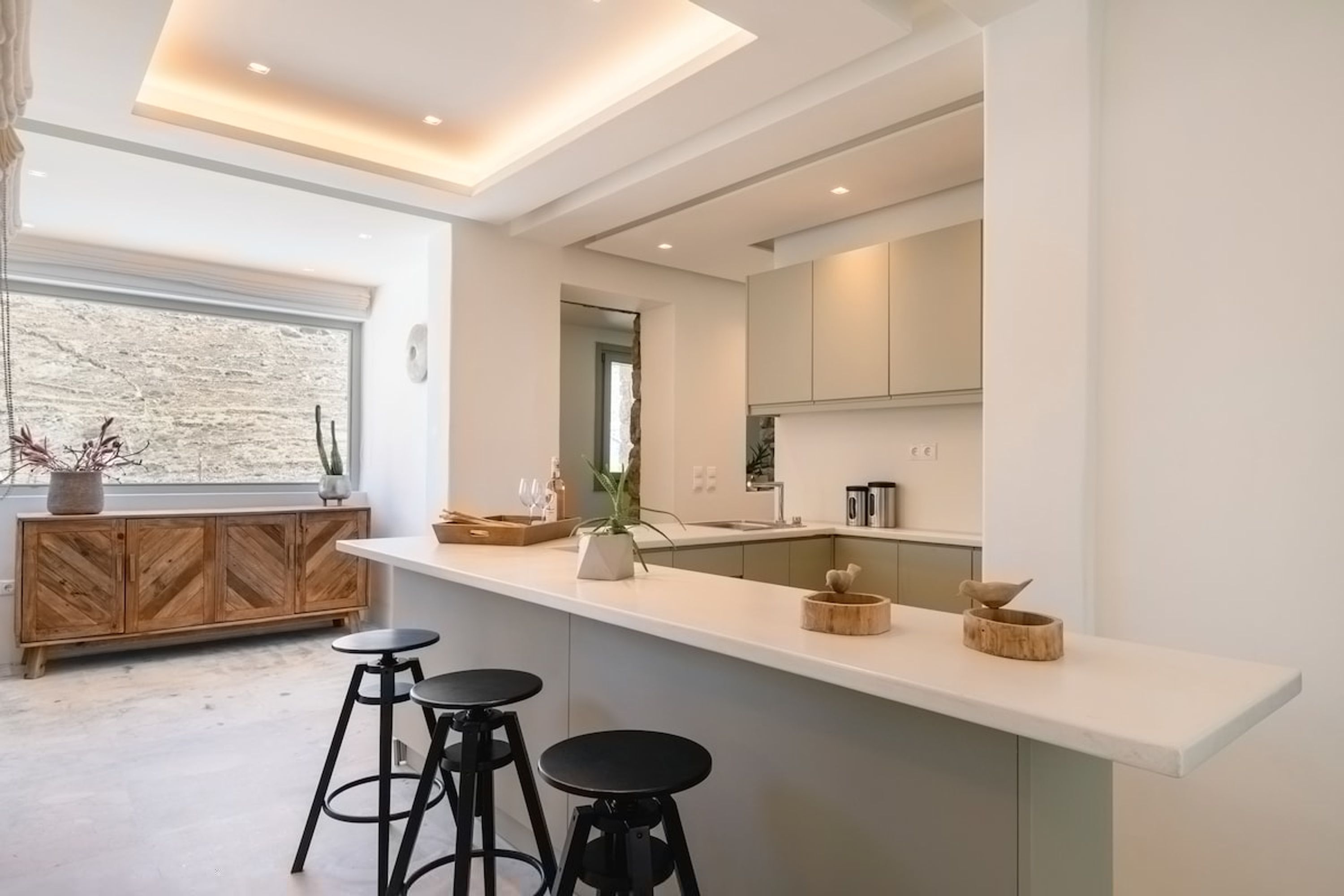 A modern kitchen with a white countertop, wooden bar stools, and a large window.