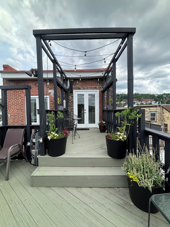 A wooden deck with a pergola, potted plants, outdoor furniture, and a view of a brick building. - Grand Brownstone with Private Rooftop & Chef's Kitchen (Pittsburgh)