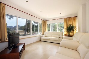A spacious living room with a large window, a flat-screen TV on a wooden stand, and a beige sofa. - Country Home "Can Medina" close to the Beach with Pool, Terrace & Garden (Cala Sant Vicenç)