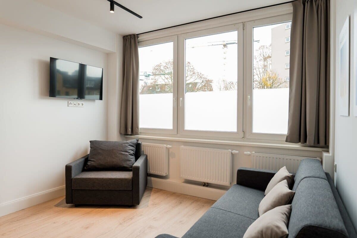 Ein modernes Wohnzimmer mit einer Couch, einem Sessel, einem Flachbildfernseher und großen Fenstern.