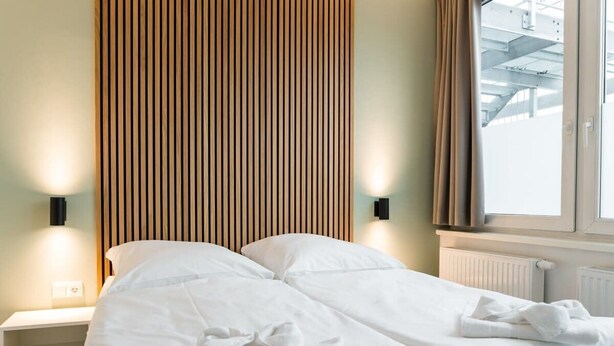 A bed with white linens, a wooden slat headboard, a nightstand with a lamp, and a window with curtains.