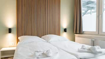 Ein Bett mit weißen Leinen, ein Kopfteil aus Holzleisten, ein Nachttisch mit Lampe und ein Fenster mit Vorhängen.