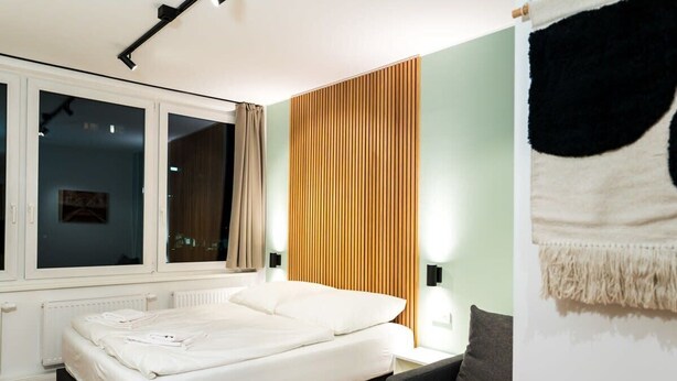A bedroom with a bed, a nightstand, a lamp, a window, and a curtain. - Standard Apartment - Homaris Apartments Hamburg Altona (Hamburg)