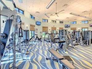 Ein Fitnessraum mit unterschiedlichen Trainingsgeräten, einem gemusterten Teppich und einem Deckenventilator.